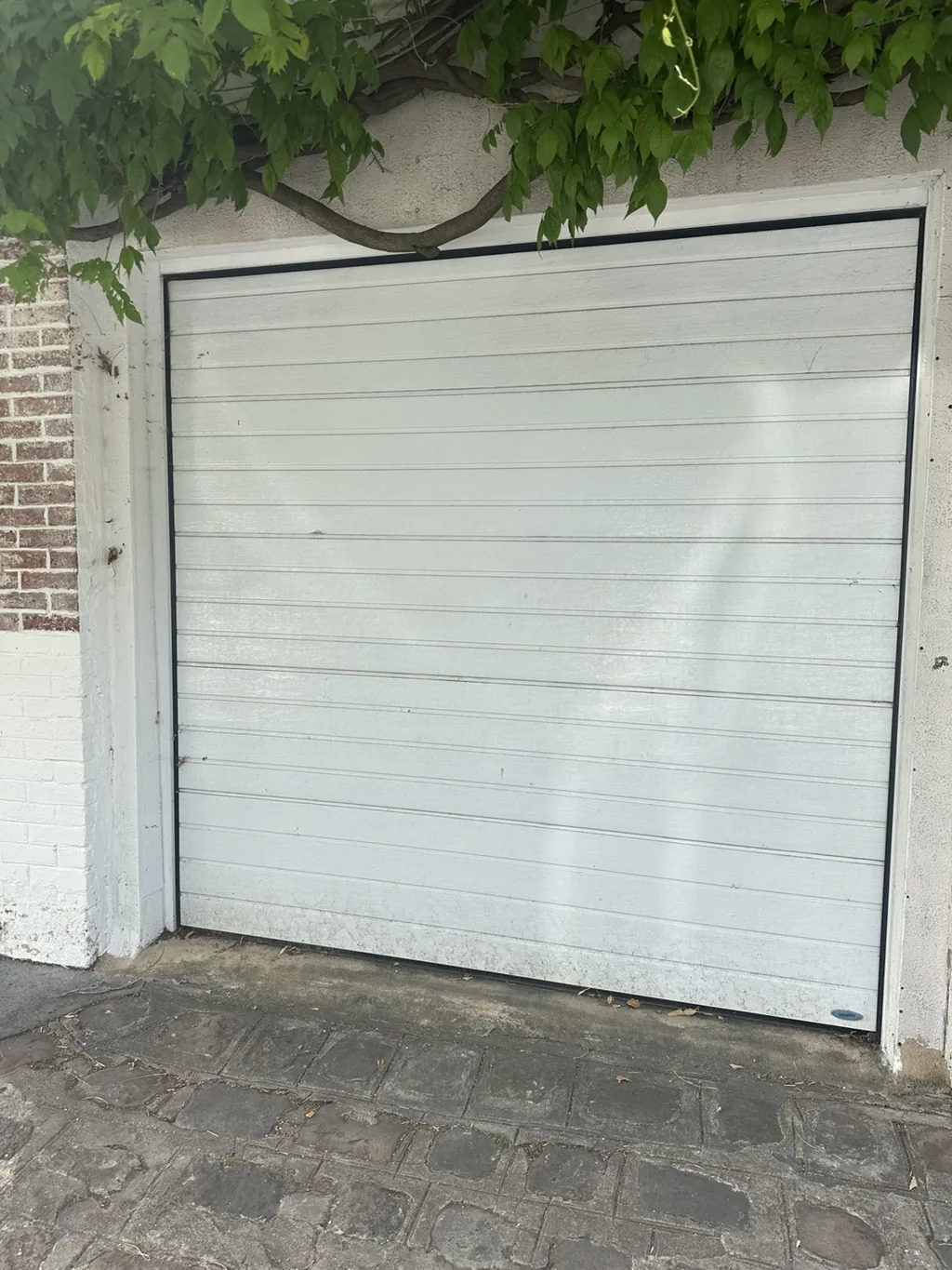 Porte de garage sectionnelle blanche installée sous une glycine, entrée pavée